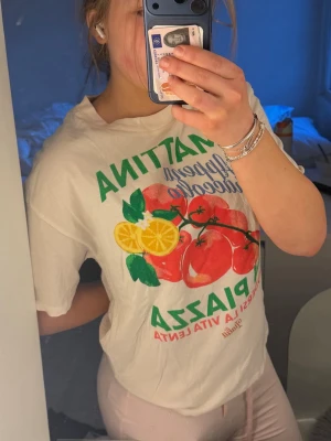 Vit t-shirt med färgglatt tryck - Säljer en vit t-shirt med stort färgglatt tryck framtill med tomater, citron och grön text. T-shirten har rund hals och korta ärmar, perfekt för en avslappnad stil. Materialet känns mjukt och bekvämt, troligen bomull.