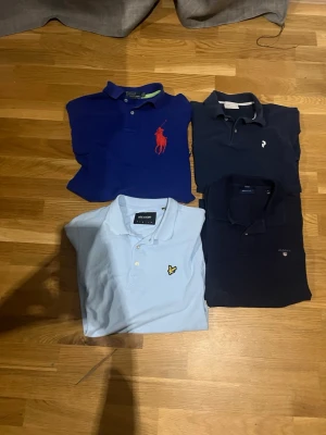 Fyra pikétröjor från Polo, Gant, Lyle & Scott - Säljer fyra pikétröjor: en blå Polo Ralph Lauren med röd logga, en mörkblå Gant, en ljusblå Lyle & Scott med gul logga och en mörkblå piké med liten vit logga. Klassiska kragar och knappar framtill. Perfekta för en stilren look.