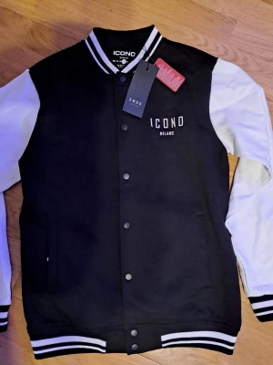 Svart och vit bomberjacka Icono Milano - Stilren svart bomberjacka från Icono Milano med vita ärmar och klassiska ränder på krage och muddar. Jackan har tryckknappar framtill och broderad logga på bröstet. Perfekt för dig som gillar sportig och trendig look.