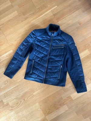 Mörkblå pufferjacka RevolutionRace - Snygg mörkblå pufferjacka från RevolutionRace med quiltade partier och flera dragkedjeförsedda fickor. Jackan har hög krage och stilrena detaljer, perfekt för kyliga dagar. Modellen heter Rocket Jacket och har en sportig look.