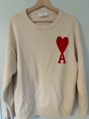 Beige stickad tröja från Ami Paris - Säljer en beige stickad tröja från Ami Paris med röd hjärtlogga och bokstaven A på bröstet. Tröjan har rund halsringning, ribbade muddar och avslut. Perfekt för dig som gillar stilrena plagg med en twist.