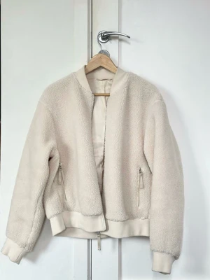 Beige teddyjacka med dragkedja - Mysig beige teddyjacka med mjukt, fluffigt material och ribbade muddar. Jackan har två fickor med dragkedja och en enkel, stilren design utan synligt märke. Perfekt för dig som gillar en clean och trendig look.