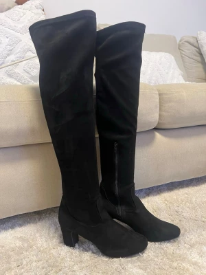 Svarta overknee stövlar med klack - Snygga svarta overknee stövlar i mockaimitation med bred klack och rundad tå. Dragkedja på insidan för enkel påtagning. Perfekta för att lyfta vilken outfit som helst och ger en trendig look.