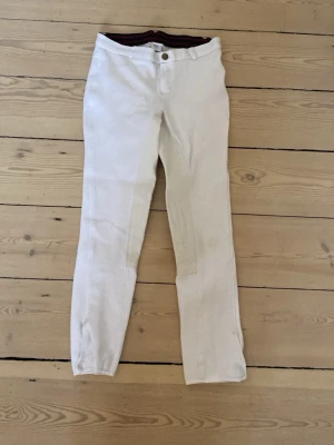 Vita byxor med helskoning - Snygga vita ridbyxor med helskoning. Byxorna har smal passform, bälteshällor och knappstängning fram. Perfekta för träning eller tävling inom ridsport. Materialet är stretchigt i midjan för bästa rörelsefrihet.