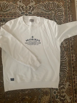 Vit sweatshirt från Morris - Vit sweatshirt från Morris med trycket 'Gentlemens Club' i mörkblått på bröstet. Tröjan har rund halsringning och ribbade muddar vid ärmslut och nederkant. Klassisk och stilren design, perfekt till jeans eller chinos. Den är storlek L men passar M