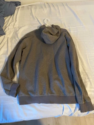 Grå hoodie från Adidas - Säljer en enkel och stilren grå hoodie från Adidas i storlek M. Hoodien har en klassisk huva och ribbade muddar vid ärmslut och nederkant. Perfekt för dig som gillar en clean och avslappnad look. Materialet är mjukt och skönt mot huden.