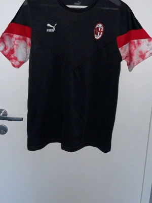 AC Milan Puma svart träningströja - Snygg svart träningströja från Puma med AC Milan-logga på bröstet. Tröjan har korta ärmar med röd och vit batikmönstrad detalj, samt Puma-logga på framsidan. Perfekt för fotbollsträning eller sportiga aktiviteter.
