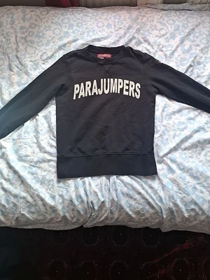 Svart sweatshirt från Parajumpers - Cool svart sweatshirt från Parajumpers med stor vit logga framtill och ett broderat märke på ärmen. Tröjan har rund halsringning, ribbade muddar och är tillverkad i mjukt bomullsmaterial. Perfekt för en avslappnad streetwear-look.