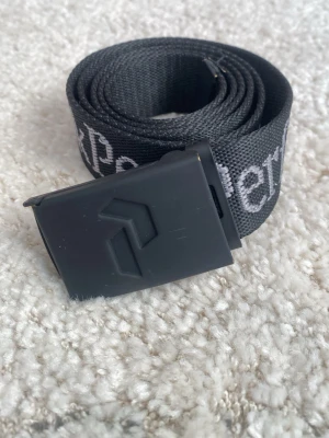 Peak Performance bälte svart - Snyggt svart bälte från Peak Performance med justerbart spänne. Bältet har texten 'Peak Performance' i vitt längs hela bandet och är gjort i slitstarkt syntetmaterial. LÄNGD: 100cm