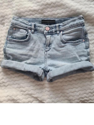 Ljusblå denimshorts från Guess - Snygga ljusblå denimshorts från Guess med uppvikta benslut och klassisk femficksdesign. Shortsen har en knapp med Guess-logga och dekorativa nitar. På ena bakfickan finns Guess-loggan i små stenar för extra detalj(några stenar har lossnat)