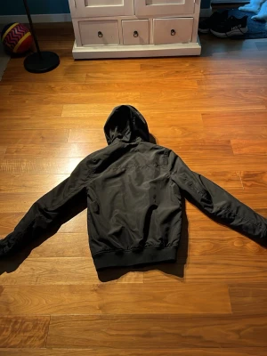Svart vindjacka med huva - Säljer en svart vindjacka med huva och dragkedja framtill. Jackan har två snedställda fickor och ribbade muddar vid ärmslut och nederkant. Perfekt för blåsiga dagar och enkel att matcha med olika outfits.