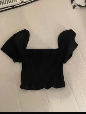 Svart croppad topp med puffärm - Trendig svart croppad topp med breda puffärmar och ribbad struktur. Perfekt för dig som vill ha en statement piece i garderoben. Snygg fyrkantig urringning och stretchigt material som sitter bekvämt.
