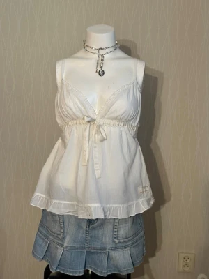 Vit linnetopp med knyt och volang - Söt vit linnetopp med smala axelband, v-ringning och knyt under bysten. Toppen har volangkant nertill och är tillverkad i ett lätt bomullstyg som känns luftigt och somrigt. Perfekt för en avslappnad och trendig look.