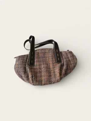 Tommy Hilfiger Handväska – Tweed / Bouclé (Unik modell) - Säljer en charmig och unik handväska från Tommy Hilfiger i vävd tweed/bouclé-stil. Väskan går i varma jordnära toner (brun/lila/beige) och har rejäla axelremmar i mörkt konstläder med snygga stickningar.  Detta är en slutsåld modell som sticker ut från mängden. Perfekt accessoar för att lyfta en enkel outfit.  Skick: Mycket fint begagnat skick!  Mått:  Bredd: 42 cm  Höjd: 24 cm  Nypris ca 1 500 kr.