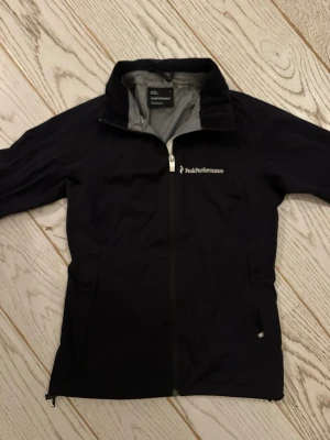 Svart vindjacka från Peak Performance - Snygg svart vindjacka från Peak Performance med hel dragkedja framtill och hög krage. Jackan har två sidofickor med dragkedja och diskret logga på bröstet. Tillverkad i lätt och slitstarkt material, perfekt för blåsiga dagar.