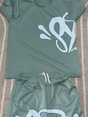Syna world grön  - Snyggt grönt set med t-shirt och shorts i mjukt material. T-shirten har ett stort vitt grafiskt tryck på framsidan och rund halsringning. Shortsen har resår och dragsko i midjan samt matchande tryck. Perfekt för en avslappnad stil.
