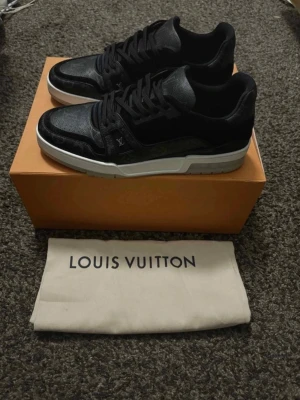 Louis Vuitton svarta sneakers - Snygga svarta Louis Vuitton sneakers med monogramdetaljer och vita sulor. Skorna har en låg profil och är tillverkade i en mix av läder och textil. Perfekta för dig som vill ha en exklusiv och trendig look.