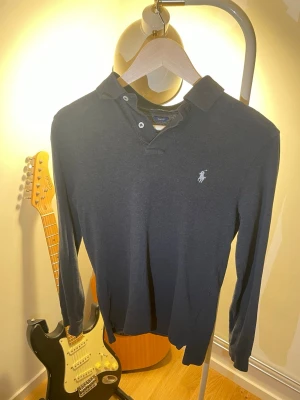 Mörkblå långärmad henley från Polo Ralph Lauren - Säljer en mörkblå långärmad henley från Polo Ralph Lauren med klassisk krage och knappar vid halsen. Ikoniska loggan broderad på bröstet. Skön och stilren skjorta i mjuk bomull, perfekt till jeans eller chinos.
