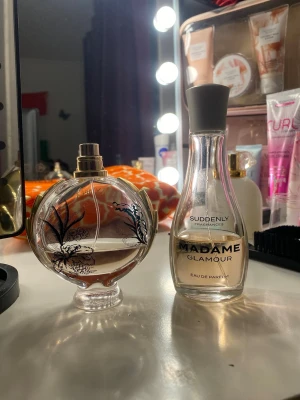 Parfymduo: Madame Glamour & Blommig flaska - Två parfymer: en Madame Glamour Eau de Parfum från Suddenly i en elegant genomskinlig glasflaska med grå kork, samt en rund glasflaska med blommigt svart mönster och gulddetaljer. Båda har en stilren och feminin design.