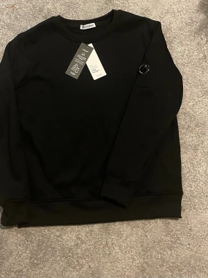 Svart sweatshirt från C.P. Company - Stilren svart sweatshirt från C.P. Company med rund halsringning och ribbade muddar. Tröjan har en ikonisk linsdetalj på ärmen och är tillverkad i mjuk bomull för maximal komfort. Perfekt för en clean och avslappnad look.