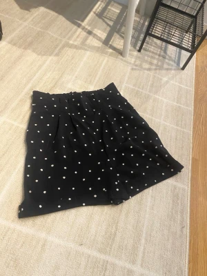 Svarta prickiga shorts - Snygga svarta shorts med vita prickar. De har en avslappnad passform och är perfekta för varma dagar. Materialet känns lätt och luftigt, vilket gör dem sköna att bära hela sommaren.