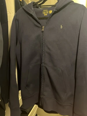 Mörkblå hoodie från Polo Ralph Lauren - Mörkblå hoodie med dragkedja från Polo Ralph Lauren. Klassisk design med huva, två fickor framtill och den ikoniska lilla loggan broderad på bröstet. Tillverkad i mjukt bomullsmaterial som är skönt att ha på sig. Perfekt för en avslappnad och stilren look.