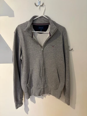 Grå zip från Gant - Klassisk grå zip hoodie från Gant med dragkedja framtill och två fickor. Liten Gant-logga broderad på bröstet och ribbade muddar vid ärmslut och nederkant. Tillverkad i mjuk bomullsblandning och har en stilren ståkrage utan huva.
