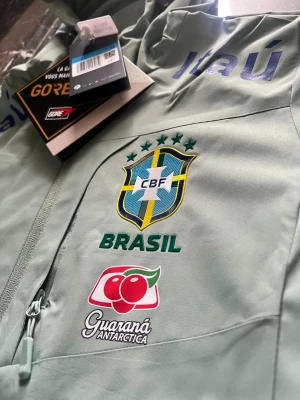 Nike Brasil GORE-TEX träningsjacka - Ljusgrön träningsjacka från Nike med Brasil-logga och flera sponsortryck som Guaraná Antarctica, Vivo och Itaú. Jackan har huva, hel dragkedja och är tillverkad i GORE-TEX-material som står emot regn och vind. Perfekt för fotbollsträning eller sportig stil.
