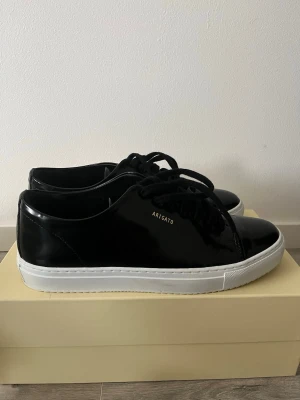 Svarta sneakers från Axel Arigato - Stilrena svarta sneakers från Axel Arigato med blank finish och vit sula. Skorna har lågt skaft, svarta skosnören och diskret guldfärgad logga på sidan. Tillverkade i läder för en clean och modern look.