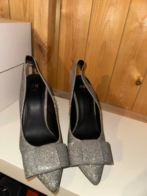 Glittriga pumps med bred klack H&M - Säljer ett par glittriga silverfärgade pumps från H&M med bred klack och öppen häl. Skorna har en spetsig tå och en dekorativ rosett framtill. Materialet är täckt av glitter som ger en riktigt festlig look. Perfekta för dig som vill sticka ut!