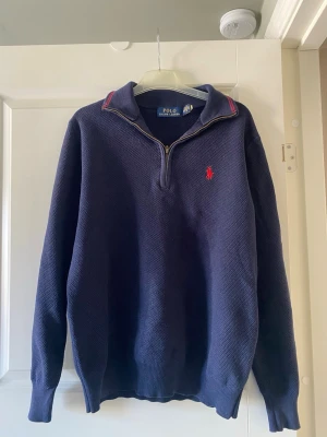 Marinblå halvzip från Polo Ralph Lauren - Säljer nu min marinblå stickad tröja från Polo Ralph Lauren med halvzip och röd broderad logga på bröstet. Tröjan har ribbade muddar, hög krage med dragkedja och diskreta röda detaljer på kragen. Perfekt för en stilren och avslappnad look. Bara att fråga om ni undrar något!