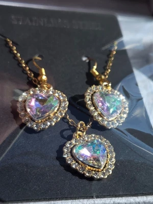 Set med hjärthalsband och örhängen - Säljer ett matchande set med halsband och örhängen i guldtonad metall. Både halsbandet och örhängena har stora hjärtformade hängen med skimrande stenar och gnistrande strasstenar runt om. Materialet är rostfritt stål och designen känns riktigt glammig och söt. Kedjan är 45 cm + förlängningskedja. Hänge är ca 1,8 - 2 cm.
