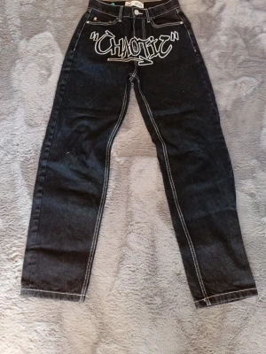 Jeans  - Säljer ett par svarta jeans med vit kontrastsöm och graffiti-inspirerat tryck 'Chaotic' framtill. Jeansen har raka ben och klassisk femficksmodell. Materialet är denim i bomull och de har en cool streetwear-vibe.