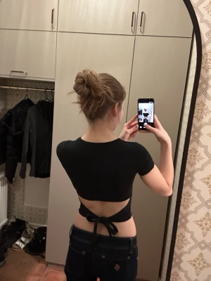 Svart croppad topp med knyt i ryggen - Snygg svart croppad topp med korta ärmar och öppen rygg som knyts baktill. Toppen är ribbad och har en tight passform som framhäver midjan. Perfekt för dig som vill ha en trendig och stilren look.