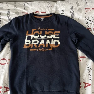 Mörkblå tröja från House - Säljer en mörkblå tröja från House med tryck 'Original House Brand Vintage' i vitt och brunt på framsidan. Tröjan har långa ärmar och en rund halsringning. Perfekt för en avslappnad stil.