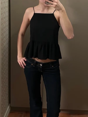 Svart volangtop med tunna axelband - Säljer en gullig svart linnetopp med tunna axelband som knyts på axlarna. Toppen har en rak halsringning och volang nedtill som ger en söt och trendig look. Perfekt att matcha med jeans eller kjol för en enkel men snygg outfit.