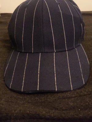 Mörkblå keps med vita ränder - Snygg mörkblå keps med tunna vita vertikala ränder i klassisk baseballmodell. Kepsen har böjd skärm och är tillverkad i ett stadigt tygmaterial. Perfekt för dig som gillar stilrena accessoarer med lite edge.