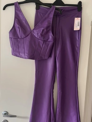 Lila set i fuskskinn med topp & byxa - Säljer ett trendigt lila set i glansigt fuskskinn. Setet består av en croppad topp med breda axelband och markerade sömmar samt ett par utsvängda byxor med hög midja. Perfekt för dig som vill sticka ut med en färgstark look.