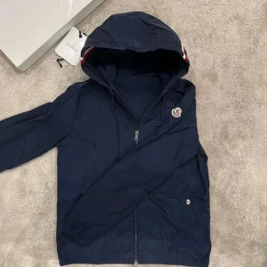 vindjacka från Moncler - Snygg marinblå vindjacka från Moncler med huva och dragkedja framtill. Jackan har Moncler-logga på ärmen och en cool rödvit rand på huvan. Lätt material i polyester, perfekt för blåsiga dagar. Äktenhets bevis kan ej garanteras. De bara hojta till om de skulle vara några frågor!