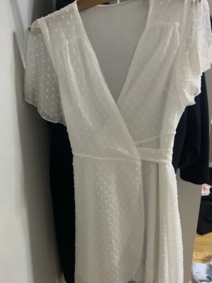 Vit prickig omlottklänning - Söt vit omlottklänning med korta, transparenta ärmar och diskret prickigt mönster i tyget. Klänningen har v-ringning och knyts i midjan för en smickrande passform. Materialet är lätt och luftigt, perfekt för varma dagar.