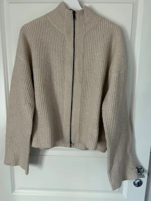 Beige stickad ziptröja med hög krage - Beige stickad tröja med dragkedja framtill och hög krage. Tröjan har en rak passform, långa ärmar och är ribbstickad för en mysig känsla. Perfekt för lager på lager under kyliga dagar.