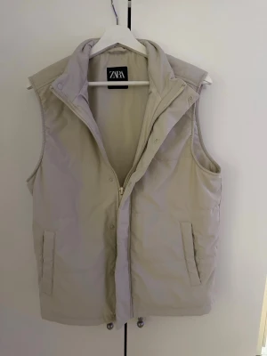 Beige dunväst från Zara - Stilren beige dunväst från Zara med hög krage och dragkedja framtill. Västen har två fickor på sidorna och en enkel, rak passform. Perfekt för lager-på-lager och ger en clean look till din outfit.