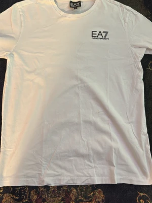 Vit EA7 Emporio Armani t-shirt - Säljer en stilren vit t-shirt från EA7 Emporio Armani med logga tryckt på bröstet. Klassisk rund halsringning och korta ärmar. Tillverkad i mjuk bomull som känns skön mot huden. Perfekt för dig som gillar enkel och snygg design.