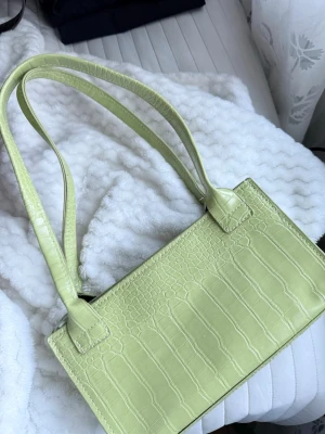 Ljusgrön axelväska i krokodil mönster  - Trendig ljusgrön handväska med krokodil mönster och raka former. Väskan har dubbla axelband och dragkedja upptill. Materialet är konstläder.