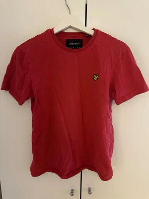 Röd t-shirt från Lyle & Scott - Snygg röd t-shirt från Lyle & Scott med klassisk rund halsringning och den ikoniska gula loggan broderad på bröstet. Tillverkad i mjuk bomull och har en normal passform med korta ärmar. Perfekt till jeans eller shorts.