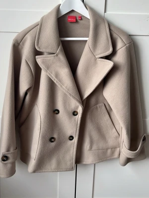 Beige dubbelknäppt kappa från RYVELS - Beige kappa från RYVLS st. 158/164. Bra skick, endast andvänd några få gånger. Stora fickor, dubbelknappar, krage kan vikas upp. Köpt på kids brandstore, nypris 700kr