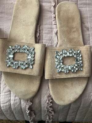 Beige sandaler med strassdetaljer - Säljer ett par snygga beige sandaler i mockaimitation med bred rem och stora, rektangulära strassdetaljer framtill. Perfekta för dig som vill ha något extra till din outfit. Mjukt material och öppen tå ger en avslappnad men glamorös känsla.