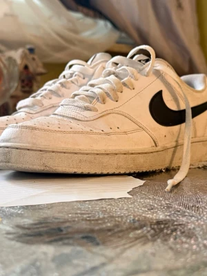 Vita Nike sneakers med svart swoosh - Säljer ett par klassiska vita Nike sneakers med svart swoosh på sidan. Skorna har lågt skaft, vita snören och perforeringar på tån för extra ventilation. Ovandelen är i läder och sulan är vit. Perfekta för en clean och sportig look.