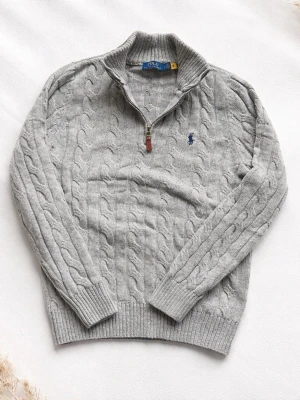 Grå kabelstickad tröja Polo Ralph Lauren - Snygg grå kabelstickad tröja från Polo Ralph Lauren med halv dragkedja och hög krage. Tröjan har det klassiska blå logobroderiet på bröstet och ribbade muddar vid ärmslut och nederkant. Perfekt för lager på lager och stilren vardagslook.