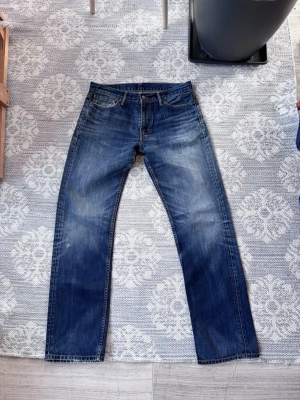 Levi's blå raka jeans - Klassiska blå jeans från Levi's med rak passform och fem fickor. Jeansen har en snygg tvättad look med subtila slitningar och kontrastsömmar. Tillverkade i slitstark denim med läderpatch bak i midjan och röd Levi's-tag på bakfickan.